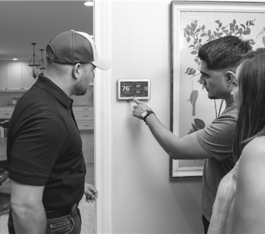 Three people stand by a wall-mounted thermostat, with one person adjusting the temperature while the others observe.