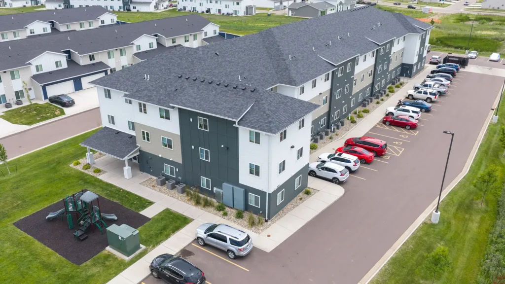 Aerial view of a modern apartment building with parked cars along one side, green lawns, a playground, and neighboring residential buildings nearby.