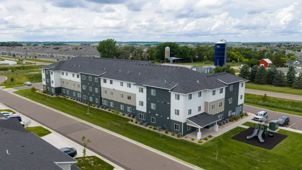A three-story apartment building with gray and white siding, surrounded by grass and sidewalks, with a small playground to the right and suburban landscape in the background.