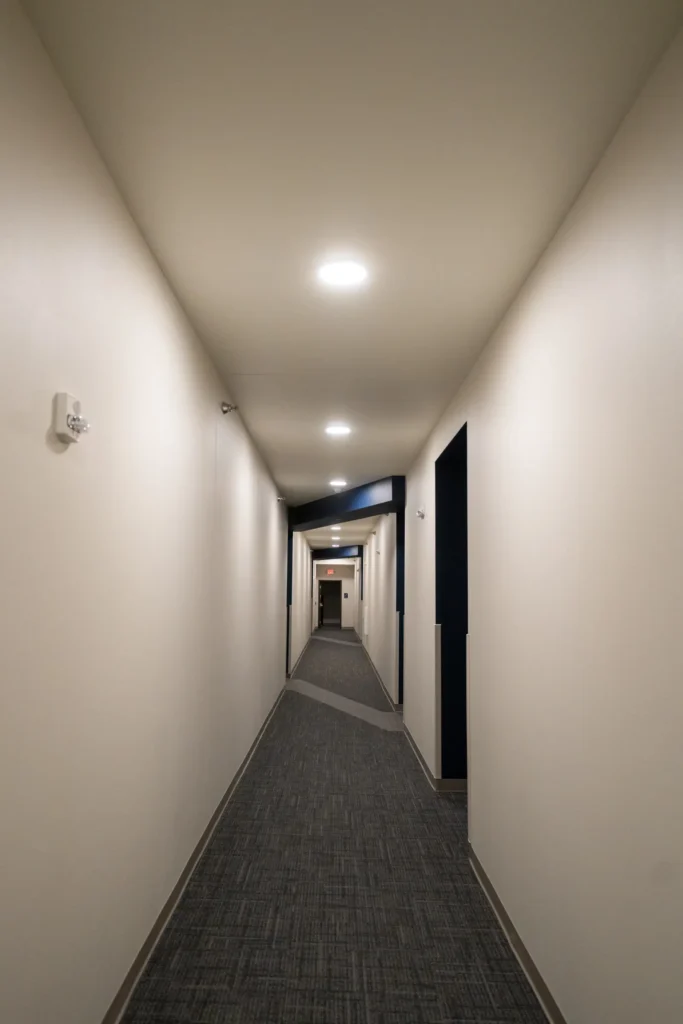 A long, well-lit hallway with white walls, gray carpet, and several doorways on the right side, extending toward a distant exit sign.