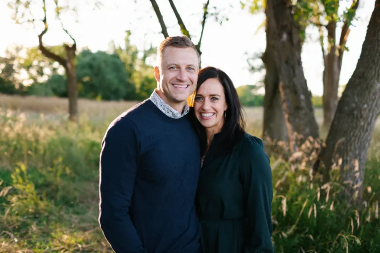 A man and woman stand close together, smiling at the camera in a sunlit outdoor setting with trees and tall grass.