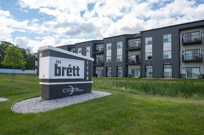 Modern apartment building with dark exterior and balconies, featuring a large sign in front that reads "The Brett" and the address "7700.