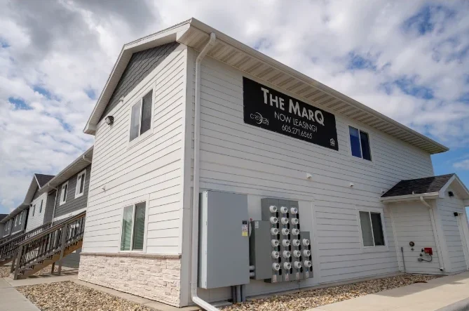 A two-story white apartment building with a sign reading "THE MARQ NOW LEASING" and several utility meters mounted on the exterior wall.